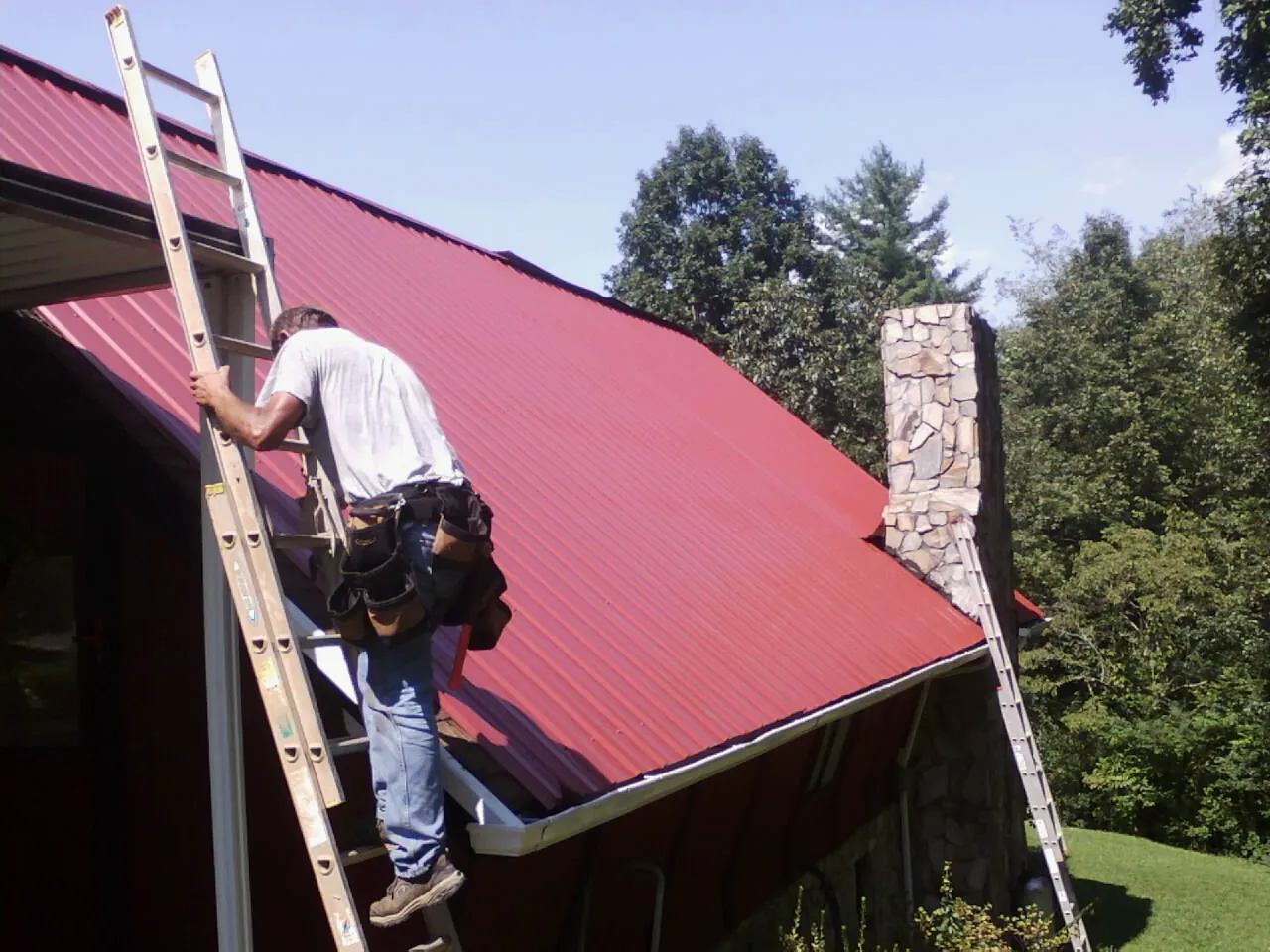 Roofer on a ladder beside a red metal roof and stone chimney — Soffit & Fascia Repair by Roofing Brothers in Monett
